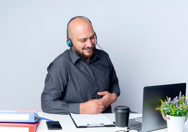 pleased-young-bald-call-center-man-wearing-headset-sitting-desk-with-work-tools-looking-laptop-putting-hand-belly-isolated-white-background_141793-84107